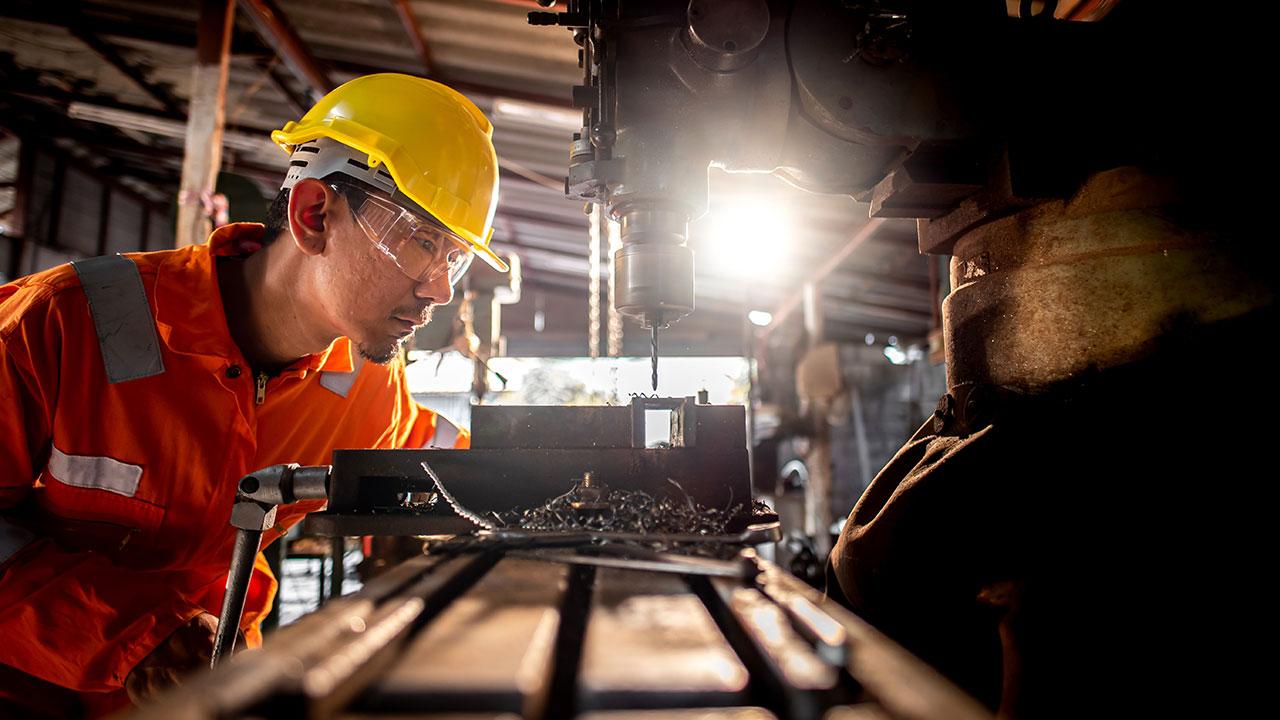 一个穿着橙色工作服，戴着眼镜的全球最大体育平台站在钻机旁边，在工业上使用金属钻头